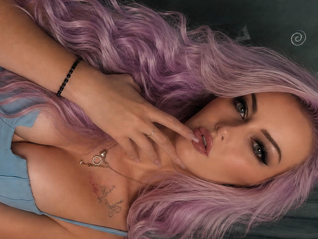 LeilahotX with pink hair, finger on lips, blue dress, tattoo on chest, green eyes, makeup, bracelet, ring, and necklace, looking seductive. Boobs, ass, tits, pussy not visible.