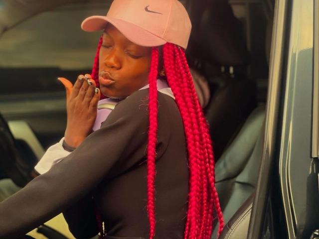 EbonyAngel, con cabello trenzado rosa y un aspecto sensual, lleva una gorra rosa Nike, una chaqueta negra y sostiene un cigarrillo, frunciendo los labios dentro de un coche. Su mano está levantada, mostrando un anillo..