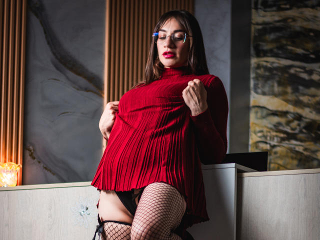 AvaStone porte un haut rouge plissé, des bas résille noirs et des sous-vêtements noirs, et elle remonte son haut pour révéler sa vulve. Elle porte des lunettes, du rouge à lèvres rouge et a de longs cheveux bruns..