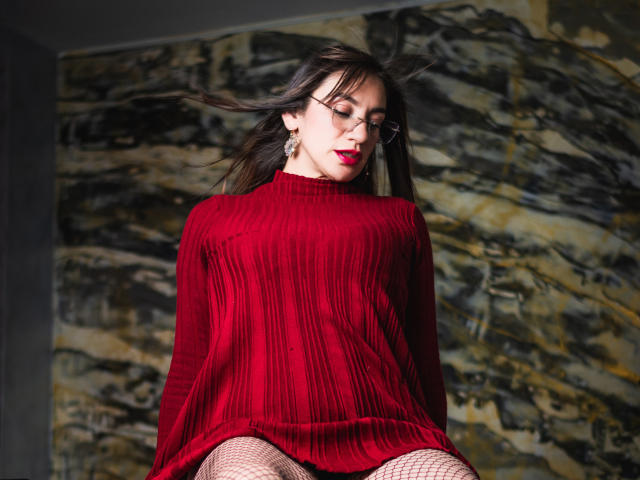 AvaStone, portant un pull rouge à rayures, des bas résille et du rouge à lèvres, avec sa poitrine légèrement visible, ses cheveux foncés flottant au vent, portant des lunettes, debout devant un mur de marbre..