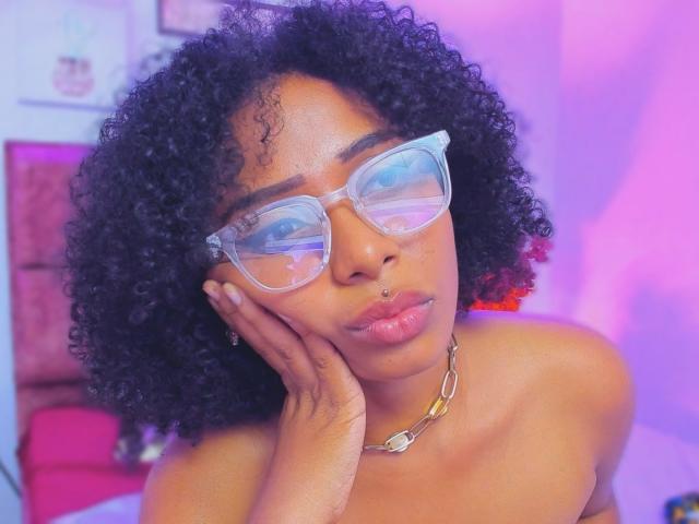 Megan Williams, com cabelo preto encaracolado, usando óculos transparentes, um piercing no nariz e um colar de corrente dourada, com a mão apoiada na bochecha. Ela está com uma aparência adorável e sexy..