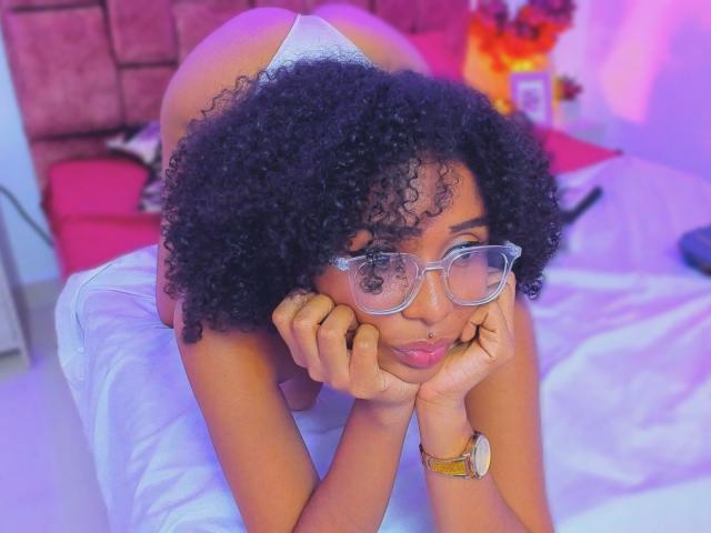MeganWilliams with curly black hair, glasses, and a nose ring, leans on a bed, hands on cheeks, wearing white underwear, exposing her ass. Purple and pink lighting.