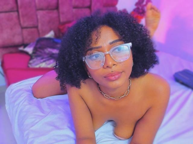 MeganWlliams with curly black hair, glasses, and a chain necklace, lying topless on a bed, her small tits and pink nipples slightly visible, looking at the camera.