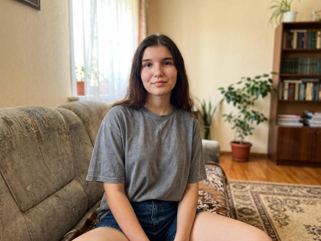 ShellaAleff with long brown hair, gray t-shirt, and denim shorts, sitting on a couch, legs apart, smiling softly, in a cozy, sunlit room.