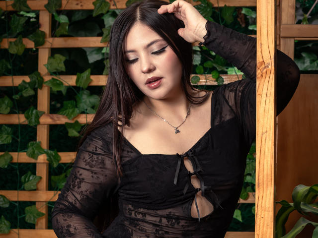 Annyek, avec ses cheveux foncés, portant un haut en dentelle noire, laissant entrevoir ses petites seins à travers une ouverture, tenant des lunettes sur sa poitrine, une main dans ses cheveux, regardant vers le bas, fond avec une feuille verte..