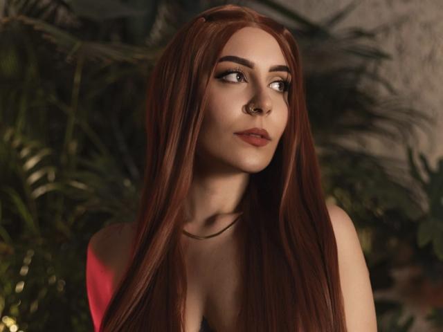 AlesziaRose with long red hair, nose ring, and subtle makeup, looking to the side with a slight smile, wearing a pink top, showing her tits.