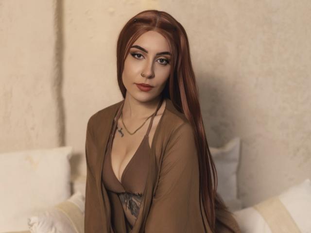 AlesziaRose has long red hair, brown eyes, and is wearing a brown bra with a sheer top, showing her small tits and a tattoo on her chest.