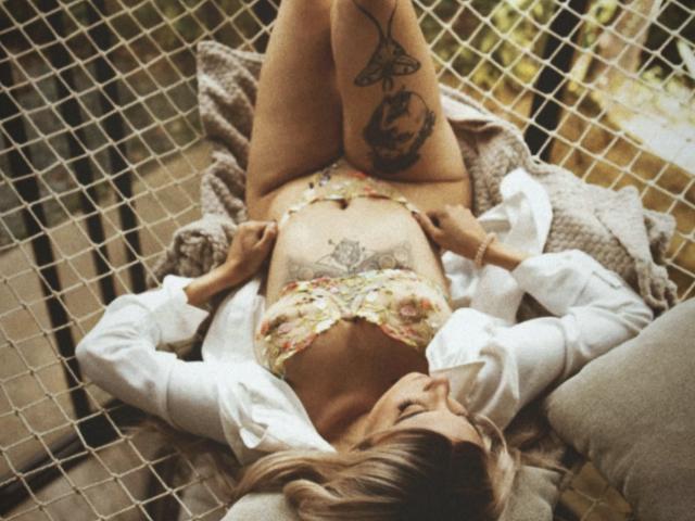 AlesziaRose, lying on a net, wearing a floral bra and unbuttoned white shirt, exposing her tits and pussy tattoo, legs up, arms on her belly, blonde hair.