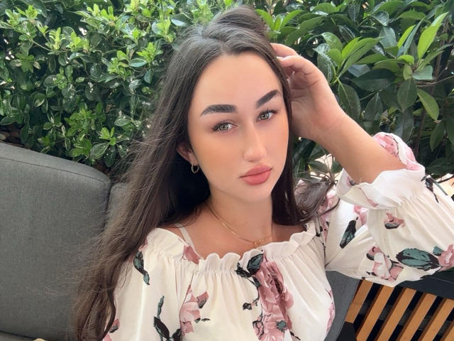 FortuneX has long dark brown hair, green eyes, and pink lips. She's wearing a floral off-shoulder top, looking at the camera with her hand in her hair.