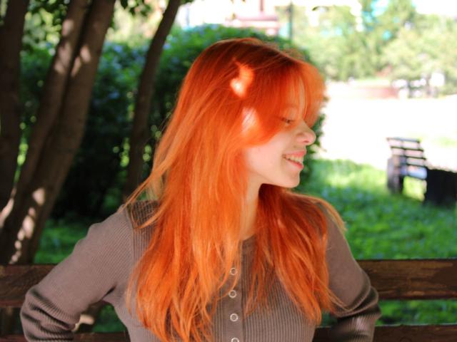 JoiMazey with bright orange hair, smiling sideways, wearing a gray ribbed shirt, hands on a wooden bench, sunlight filtering through trees, green grass in the background.