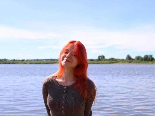 JoiMazey with red hair, smiling, eyes closed, wearing a brown button-up shirt, standing in a blue river, sunlight on her face, gentle water ripples.