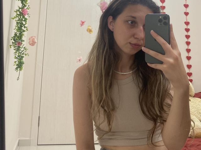 Sanella with long brown hair, small tits, wearing a beige crop top, holding a phone, taking a mirror selfie, looking slightly to the side, in a white room.