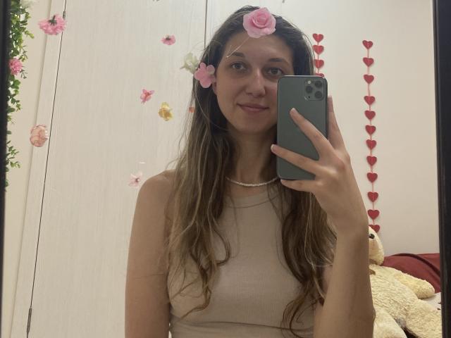 Sanella with long brown hair, pink flower headpiece, beige top, holding green phone, smiling, white wall, red heart decals, beige teddy bear, pearl necklace, cute, mirror selfie.