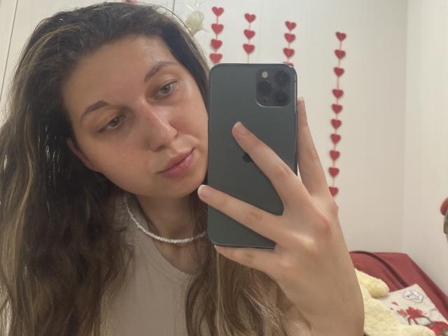 Sanella's selfie shows her with wavy brown hair, light skin, and a pearl necklace, holding a green phone, wearing a white shirt, with a subtle smile.