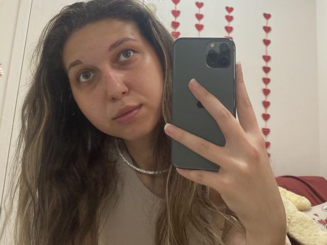 Sanella with long brown hair, holding a black phone, wearing a white top and pearl necklace, taking a selfie, showing her tits slightly in the mirror.