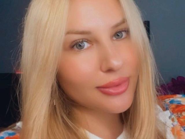 AvaQueen with blonde hair, blue eyes, and pink lips, has smooth fair skin and a slight smile, wearing a colorful floral top. Her tits are not visible.