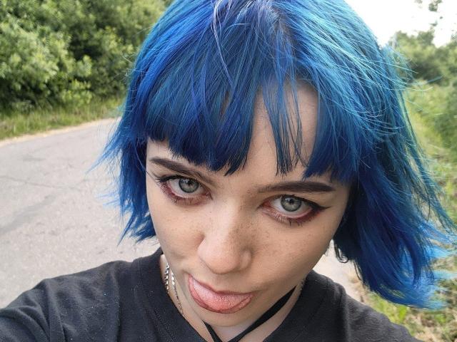 ParisDemirjian with blue hair, pouty lips, and green eyes, wearing black shirt, has a chain necklace, and raised eyebrows, looking directly at the camera.