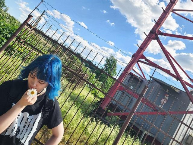 ParisDemirjian with blue hair, black shirt, holding daisy, standing by rusty fence, red metal structure, and gray building, under blue sky with white clouds.