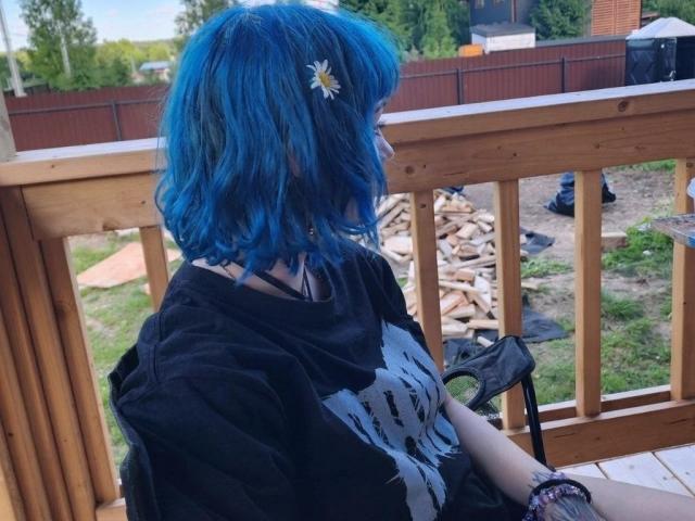 ParisDemirjian with blue hair, daisy hair clip, black shirt, and black choker, sitting on wooden porch, holding black glove, looking outside, visible wooden railing and yard.