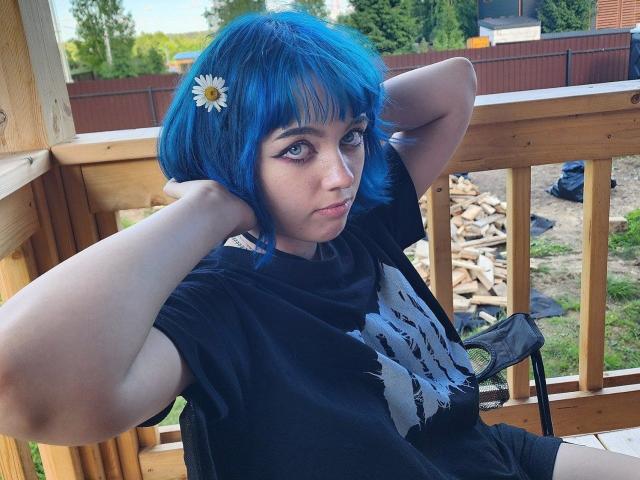 ParisDemirjian with blue hair, daisy in hair, black tee, arms behind head, looking at camera, on wooden balcony, outdoor, daytime, standing, hands on head, short hair, serious expression.
