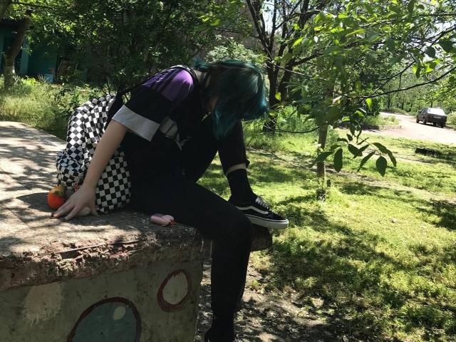 ParisDemirjian with teal hair, black shirt, checkered backpack, black pants, and sneakers, sitting on a concrete ledge, holding a pink phone, with an apple beside her.