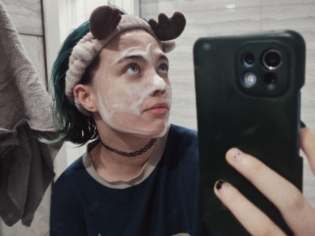 ParisDemirjian with a white face mask, wearing a brown reindeer headband, green hair, black choker, and navy shirt, taking a mirror selfie with a green phone.
