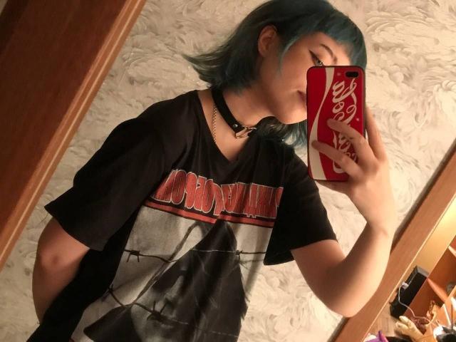 ParisDemirjian with blue hair, wearing a black "Oasis" t-shirt, black choker, and holding a red iPhone with a Coke logo, taking a mirror selfie.