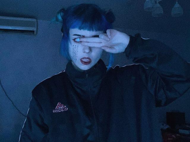 ParisDemirjian with blue hair in double buns, black Adidas jacket, covering one eye with her hand, dark lipstick, white face markings, looking surprised, in blue-tinted photo.