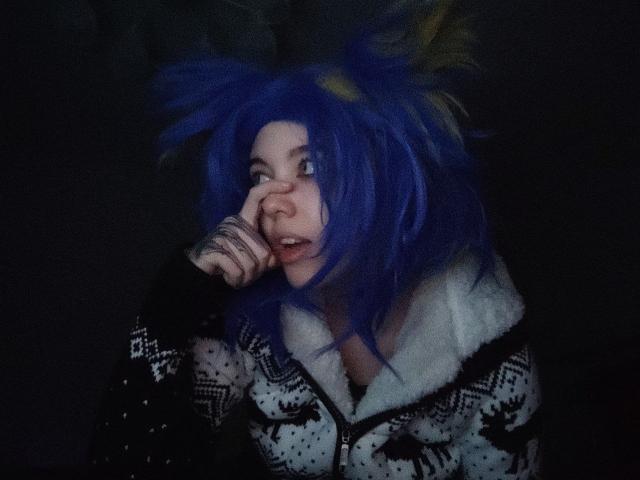 ParisDemirjian with blue hair, white furry jacket, black pattern, and surprised expression, resting chin on hand, looking to the side. Dark background.