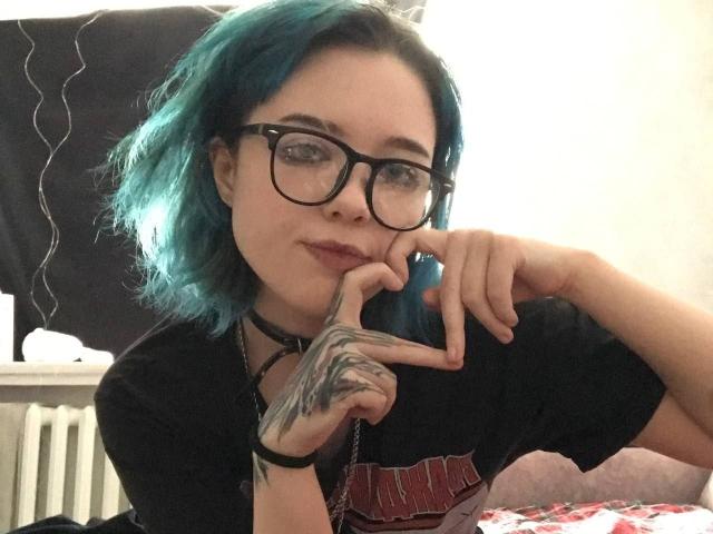 ParisDemirjian with blue hair, black glasses, and tattoos on her right hand, resting her chin on her fingers, wearing a black shirt and choker, looking slightly smug.