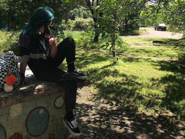 ParisDemirjian with teal hair, black shirt, black pants, and black sneakers, sitting on a graffiti-covered wall, eating a lollipop, with a checkered bag and a red toy on left.