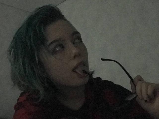 ParisDemirjian with green-tipped hair, eating with a spoon, holding glasses, wearing a red plaid shirt, looking to the side, dark, moody lighting, slight blush on her face.