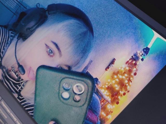 ParisDemirjian with blue eyes, wearing headphones, striped shirt, holding green phone, selfie with city lights in background.