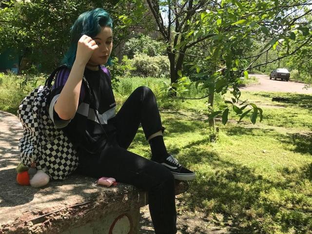 ParisDemirjian with teal hair, black outfit, and checkered bag, sitting on a stone, raising hand, green grass, and trees, black sneakers, pink toy, and orange ball nearby.
