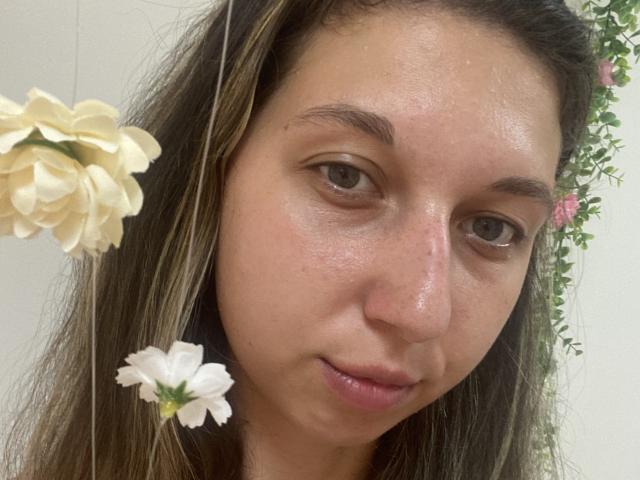 Sanella's face with slightly shiny skin, brown eyes, and light brown hair, wearing two white and beige flowers on her left side. She's looking at the camera with a subtle smile.