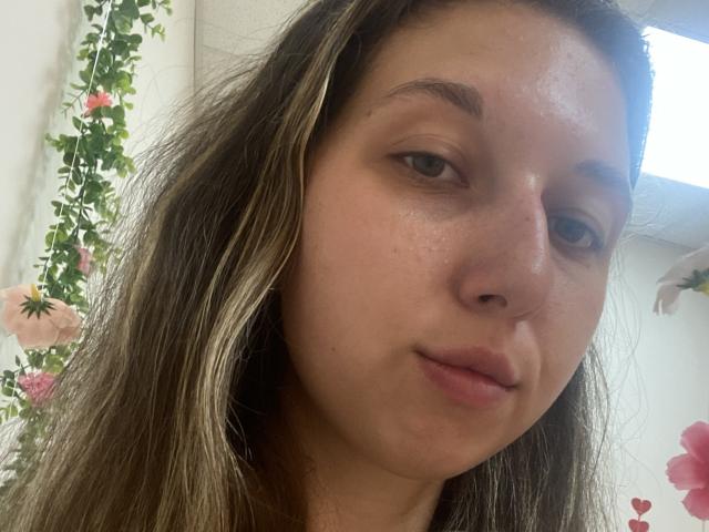 Sanella's face with slightly parted lips, brown eyes, and wavy brown hair, showing a relaxed, almost smug expression, with a subtle sheen on her skin.