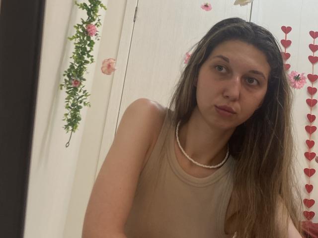 Sanella has long brown hair, a pearl necklace, and a beige sleeveless top, with a neutral expression, in a slightly angled selfie, showing her tits and ass.