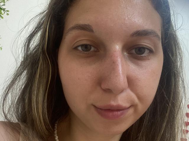 Sanella's face, brown eyes, slight smile, light brown wavy hair, fair skin, subtle freckles, no makeup, wearing a white bead necklace, slightly shiny forehead.