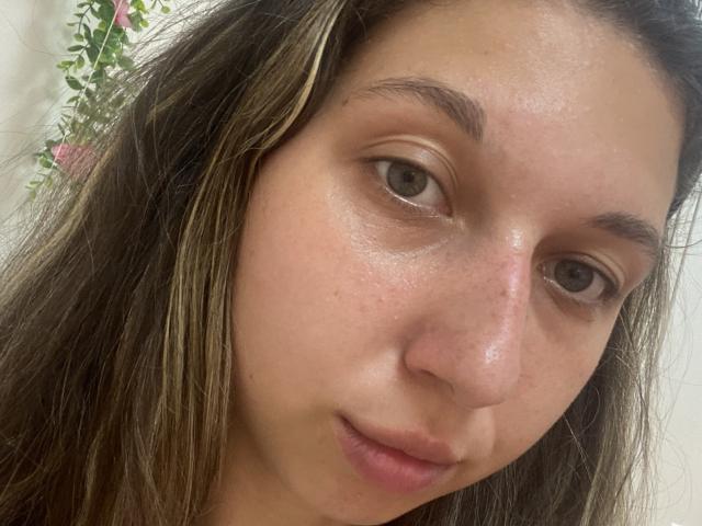 Sanella's face with light brown skin, brown eyes, and long brown hair with blonde highlights. She has a slight smile, visible nose, and subtle freckles on her cheeks.