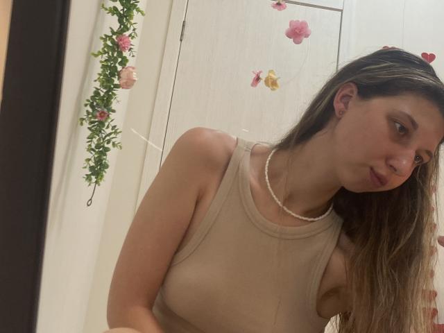 Sanella with long brown hair, wearing a beige tank top, showing her small tits, and a pearl necklace, leaning forward in a mirror selfie.