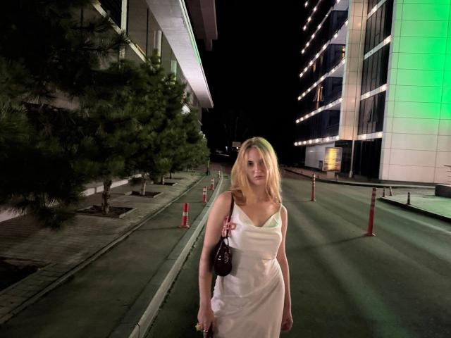 Viloriia, com cabelo loiro, vestido branco e bolsa marrom, está em uma rua vazia à noite, com uma expressão séria, segurando a parte superior do vestido, com um prédio com luzes verdes ao fundo..