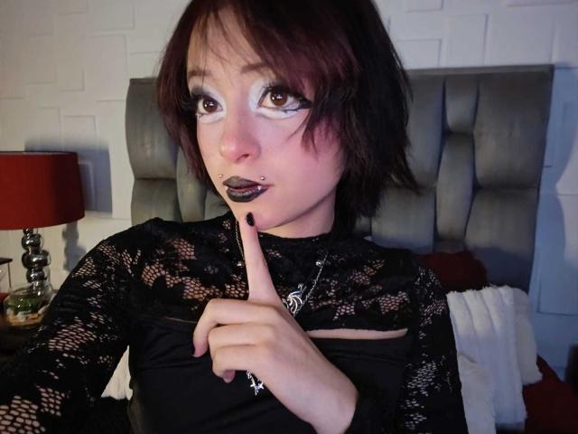 ViolettClay, avec des cheveux roux foncés, un haut en dentelle noire, du rouge à lèvres noir, un piercing au nez, un doigt sur les lèvres, tenant une lampe en arrière-plan, assise sur un lit avec une tête de lit grise..