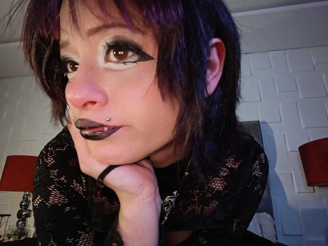 ViolettClay with dark purple hair, black lipstick, and nose ring, wearing a black lace top, resting chin on hand, brown eyes with dramatic eyeliner.