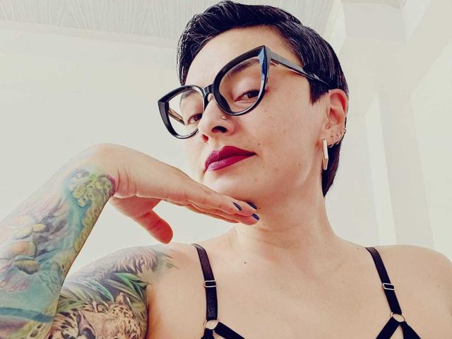 KatParadise, con i capelli neri corti, gli occhiali neri, il rossetto rosso, tatuaggi sul braccio destro e un reggiseno nero, che si tocca il mento con una mano, sorridendo..