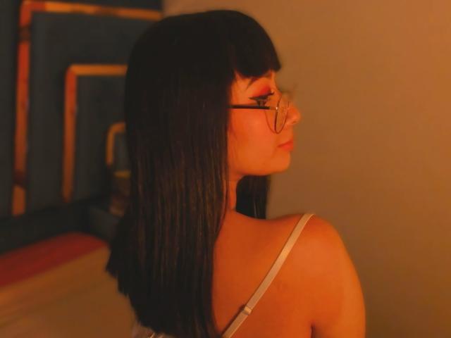 Genesiss with straight black hair, wearing glasses and a white strap top, showing her back and a subtle sideboob, in warm orange lighting.