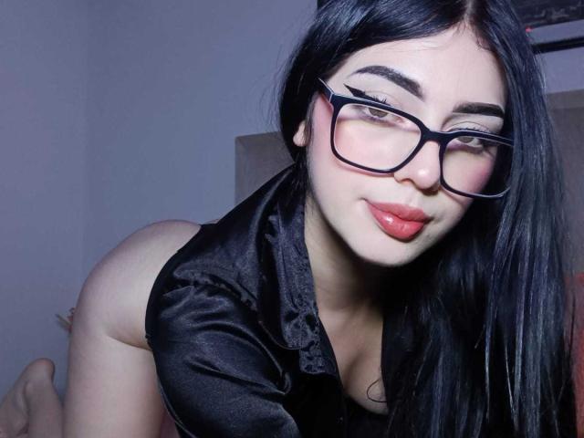 Isabella Rossi mit schwarzen Haaren, Brille, rotem Lippenstift und einem schwarzen Satin-Top, die nach vorne gebeugt ist, ihren Hintern zeigt und lächelt. Ihre Brüste sind klein, ihr Hintern ist rund..