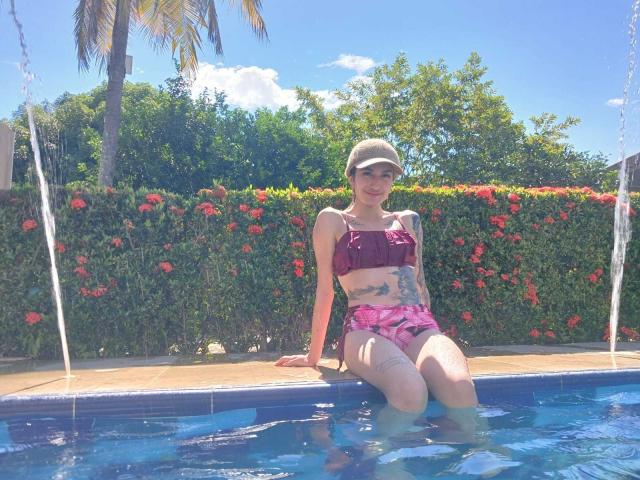 Kaiix has small tits, wears a magenta bra and pink shorts, sitting in a pool with water up to her thighs, wearing a beige cap, tattoos on her stomach.