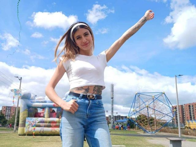 Kaiix con tatuajes, camiseta blanca ajustada, jeans azules, cinturón con hebilla dorada con la letra "G", brazo levantado, sonriendo, cabello rubio, cielo azul, nubes blancas y estructuras inflables en el parque..