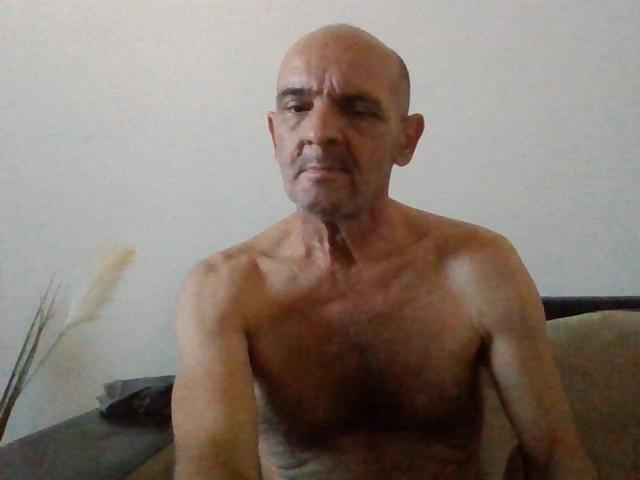 SkorpioCumShower senza camicia, calvo, con un'espressione burbera, petto cadente e un corpo leggermente rugoso e abbronzato, è seduto su un divano con un'aria pensierosa..