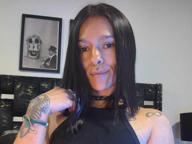 Amelie Blake, con capelli neri, piercing al naso e collana, sorridente, mostra tatuaggi sul braccio destro e sulla spalla sinistra, indossa un top nero con spalline, in una camera da letto. In secondo piano, un ritratto di Skelly..